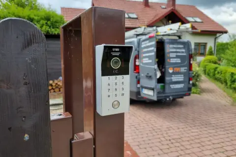 Nowy domofon na furtce. Zastanawiasz się, ile kosztuje montaż domofonu w domu jednorodzinnym? W tle widać dom i samochód firmy instalacyjnej.