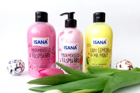 Trzy butelki ISANA: "Mademoiselle Raspberry" (różowa, z pompką i bez) i "Lady Lemon & Mr. Mint" (żółta). Obok kwiaty i ozdobne kule.