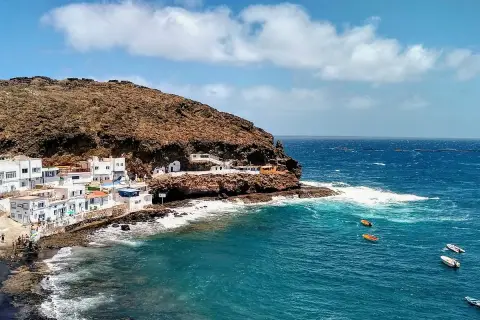 Pueblo costero blanco anidado en la ladera rocosa de **Tufia**. El mar azul turquesa choca contra las rocas, con pequeñas embarcaciones flotando.