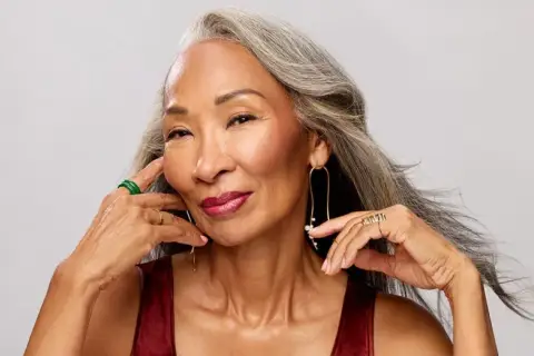 Mujer con cabello gris, con un elegante efecto de grey blending, sonríe con labios rojos y joyas doradas y verdes.