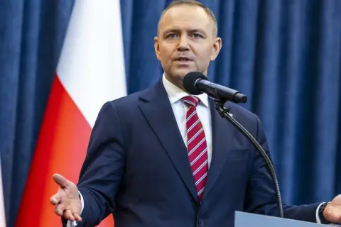 Mężczyzna w garniturze przemawia przy mikrofonie na tle flagi Polski. Czy prezydent faktycznie planuje ogłosić stan wyjątkowy w związku z sytuacją na wschodzie?