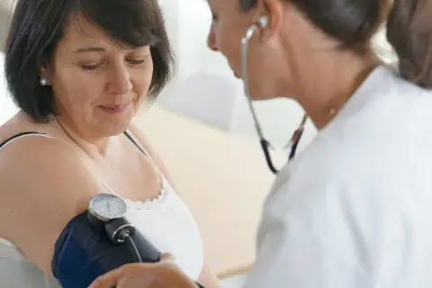 Lekarka bada ciśnienie pacjentce. Na ramieniu kobiety widoczny jest mankiet ciśnieniomierza, a lekarz słucha stetoskopem.