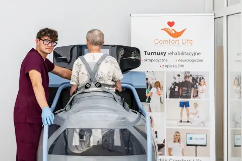 Rehabilitacja po operacji kręgosłupa: pacjent w specjalistycznym urządzeniu, asystuje mu terapeuta. Na banerze informacje o turnusach.