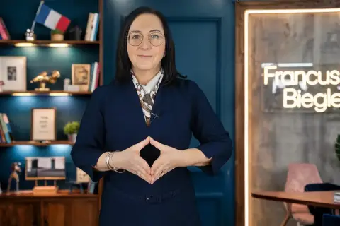 Kobieta w okularach i granatowej sukience, z rękami złożonymi w romb, na tle regału z książkami i neonu z napisem "Francuski Biegle".