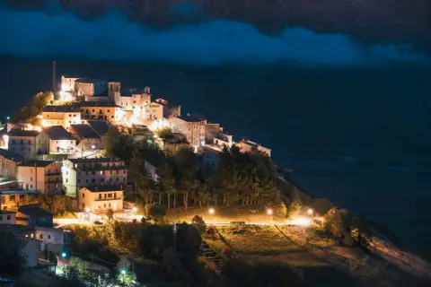 Norcia: cosa vedere dopo il terremoto. Borgo illuminato di notte, arroccato su un colle, con case in pietra e alberi.