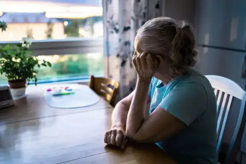 Starsza kobieta siedzi przy stole, zmartwiona. Zima i odcięcie prądu sprawiają, że czuje się samotna i bezradna.