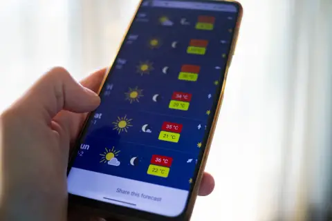 Dłoń trzyma smartfon z aplikacją pogoda, pokazującą wysokie temperatury i słoneczną aurę.