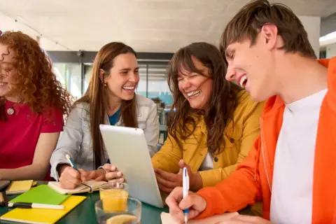 Uśmiechnięci studenci, którzy uczą się psychologii na studiach zaocznych, pracują razem przy stole, korzystając z laptopa i tabletu.