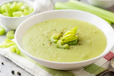 Zuppa cremosa di sedano verde, guarnita con gambi freschi e pepe nero. Accanto, una ciotola di sedano a cubetti.