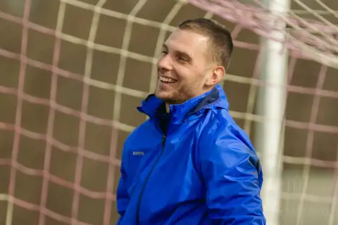 Uśmiechnięty piłkarz w niebieskiej kurtce, gotowy do gry w stylu tiki-taka.
