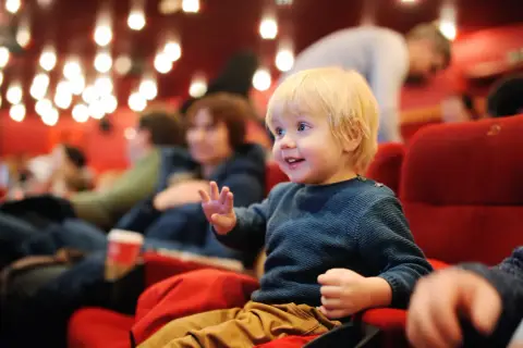 Mały chłopiec z blond włosami siedzi w kinie, ciesząc się filmem. Czy w kinie sprawdzają wiek dziecka?