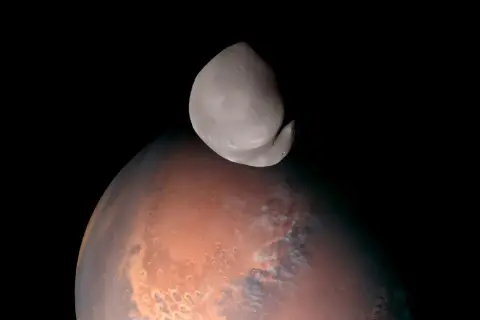Niewielki, nieregularny księżyc Deimos unosi się nad czerwoną planetą Mars.