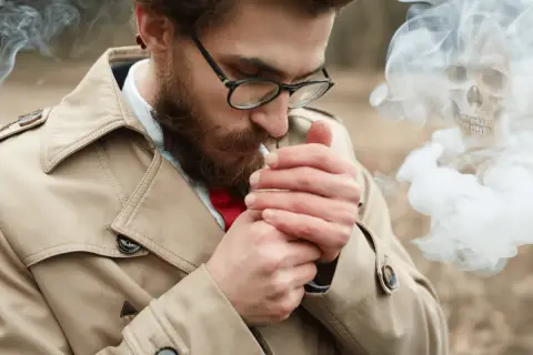 Hombre con barba enciende cigarrillo. El humo forma una calavera, advirtiendo sobre la alergia al humo del tabaco.