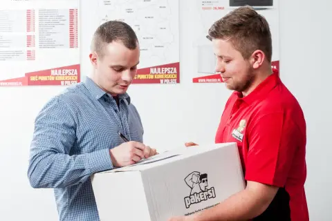 Listonosz w czerwonej koszulce przekazuje paczkę mężczyźnie w kratę, który podpisuje dokument.