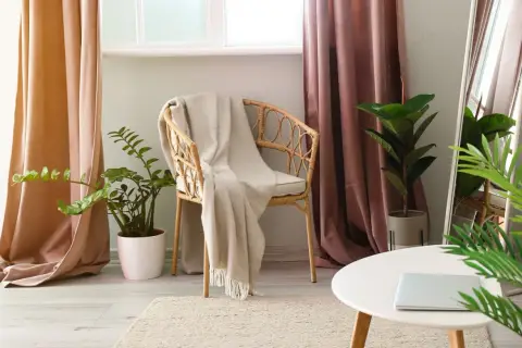 A cozy reading nook with a wicker chair draped in a blanket, flanked by pooled curtains in soft peach and dusty rose hues.