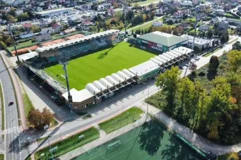 Widok z lotu ptaka na stadion Górnik Łęczna z zieloną murawą, trybunami i otaczającymi go budynkami.