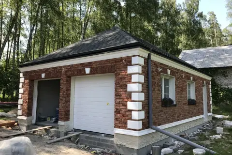 Nowy garaż z cegły, z białym gzymsem i ozdobnymi pilastrami, otoczony zielenią.