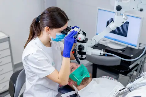 Dentystka w masce i rękawiczkach bada ząb pacjenta pod mikroskopem. Na ekranie widać zdjęcie rentgenowskie. To nowoczesne leczenie kanałowe, cena zabiegu zależy od skomplikowania.