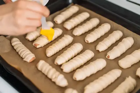 Ręka smaruje pędzelkiem parówki zawinięte w ciasto, przygotowując je do pieczenia. To prosty przepis na parowance.