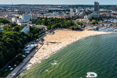 Słoneczny bulwar nadmorski Gdynia tętni życiem. Plaża pełna ludzi, a w oddali port z żaglówkami i miejska panorama.