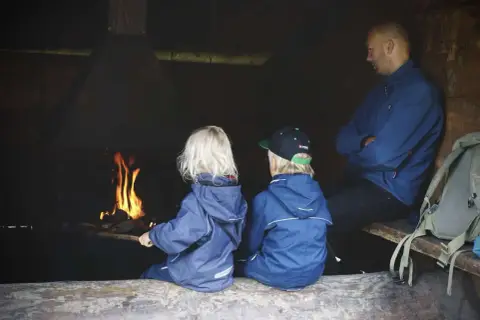 Två barn och en vuxen sitter vid en eld. En mysig paus under vandring med barn.