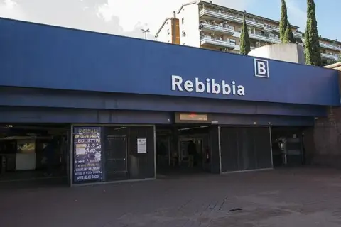 Ingresso della metro Rebibbia a Roma, con insegna blu e lettere bianche. Edifici residenziali sullo sfondo.