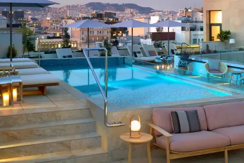 Piscina en la azotea del Barceló Raval con vistas a la ciudad al atardecer. Tumbonas, sofás y luces crean un ambiente acogedor.