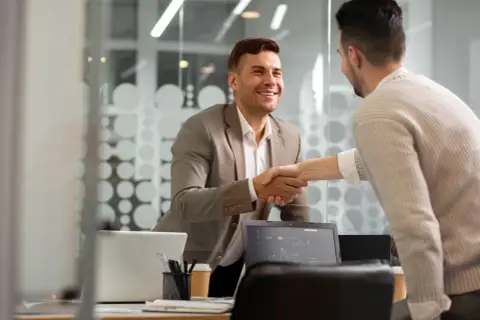 Dwóch mężczyzn w biurze ściska sobie dłonie. Jeden w garniturze, drugi w swetrze. Rozmowa o tym, resell co to, zakończona sukcesem.