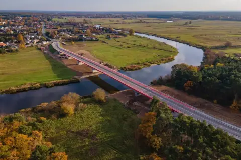 Nowy, czerwony most na warcie przecina spokojną rzekę, łącząc malowniczą wieś z zielonymi polami i lasami jesienią.