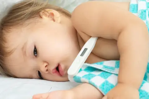 Małe dziecko leży pod kocykiem, a pod pachą ma termometr dla niemowlaka.