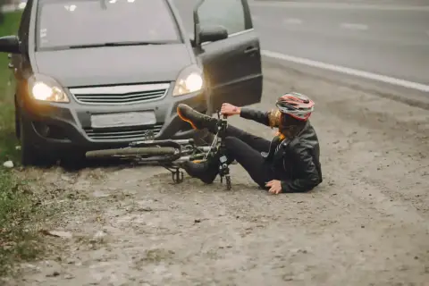 Kobieta w kasku leży obok przewróconego roweru, obok stoi samochód z otwartymi drzwiami. Wygląda na to, że doszło do ucieczki z miejsca kolizji.