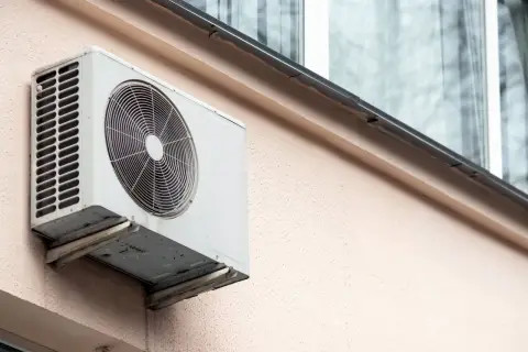 Klimatyzacja w bloku, biała jednostka zewnętrzna zamontowana na jasnoróżowej ścianie, z widocznym wentylatorem i kratką.