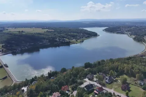 Widok z lotu ptaka na zalew Brody, otoczony lasami i domami. Widać tamę i fragment torów kolejowych.