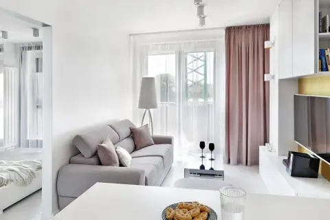 Jasny, bardzo mały salon w bloku z szarą kanapą, stolikiem kawowym i widokiem na balkon. Obok widać łóżko.