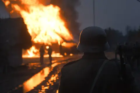 Żołnierz w hełmie obserwuje płonące chaty w świetle dnia. W tle widać innych żołnierzy i druty kolczaste.