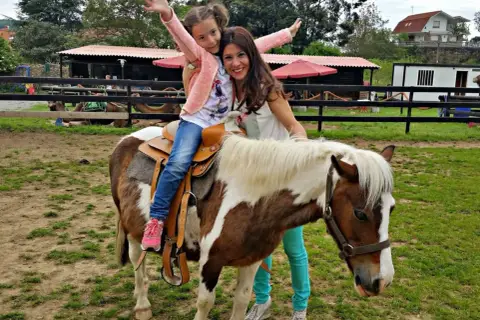 Niña feliz en un poni, disfrutando de una ruta a caballo en Cantabria. Madre sonriente la acompaña.
