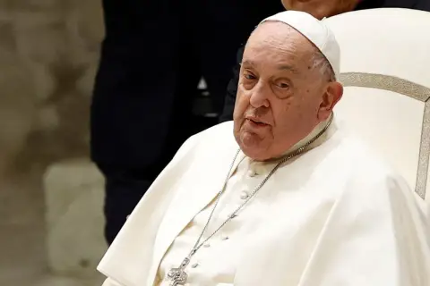 Papież Franciszek, siedzący na białym krześle, w białej sutannie i piusce. Jego wzrost jest typowy dla mężczyzny w jego wieku.