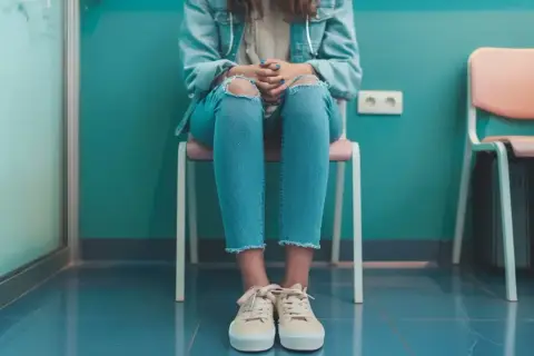 Dziewczyna w podartych dżinsach siedzi na krześle, czekając na wizytę. Jej dłonie splecione na kolanach mogą sugerować niepokój lub pochwicę.