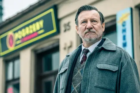 Mężczyzna w eleganckim swetrze i płaszczu, z siwą brodą, stoi przed budynkiem z neonem. To jeden z aktorów z obsady "Minuta ciszy".