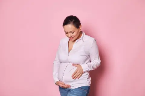 Uśmiechnięta kobieta w ciąży po 40. delikatnie dotyka brzucha na różowym tle.