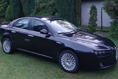 Czarna Alfa Romeo 159 lśni na trawie, otoczona zielenią drzew.