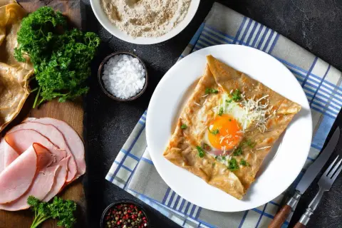 Francuska crepe z jajkiem sadzonym, serem i natką pietruszki na talerzu, obok szynka i składniki.