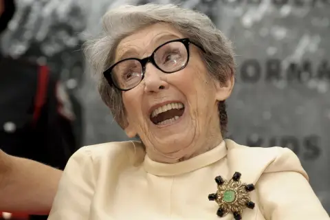 Uśmiechnięta babcia po francusku, w okularach i eleganckiej broszce, z otwartymi ustami.