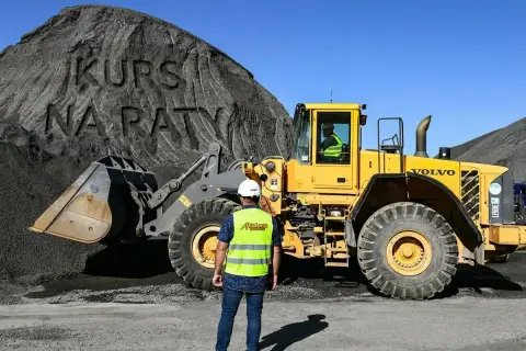 Pracownik w kamizelce odblaskowej obserwuje ładowarkę kołową. Na hałdzie widnieje napis "KURS NA RATY", sugerujący szkolenie z obsługi maszyn.