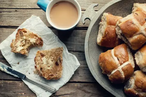 Na drewnianym stole leżą świeże, pachnące hot cross buns, obok kubek kawy i kromka posmarowana masłem.