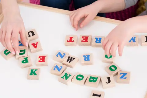Dzieci układają litery na tablicy, ćwicząc podział na głoski. Widoczne litery to m.in. U, A, T, R, E, N, K, P, Z, O, B, J, I.