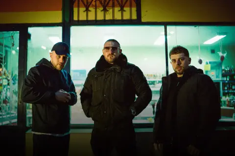 Trzech mężczyzn w ciemnych kurtkach, tworzących zgraną hałastrę, pozuje przed oświetloną witryną sklepową.