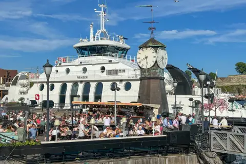 Tętniący życiem deptak w Oslo, z ludźmi siedzącymi w kawiarniach, na tle białego statku i zabytkowej wieży zegarowej. Oslo czy warto? Zdecydowanie tak!