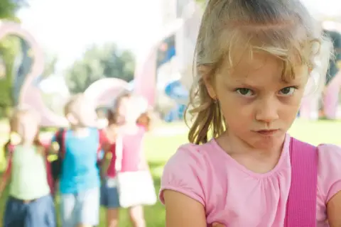 Zagniewana dziewczynka, bunt 7 latka, na tle bawiących się dzieci i placu zabaw.