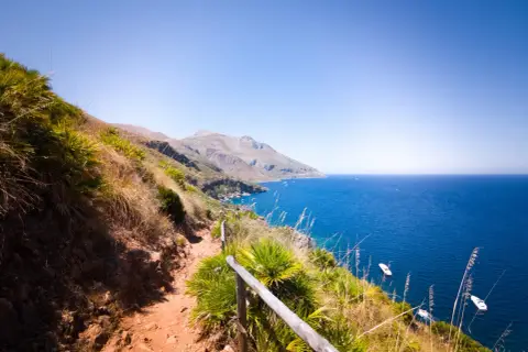 A scenic hiking trail winds through the rugged landscape of Lo Zingaro Nature Reserve, overlooking the brilliant blue Mediterranean Sea dotted with boats.